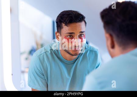 Uomo biraciale sorridente con maschere sotto gli occhi che guardano in specchio in bagno Foto Stock