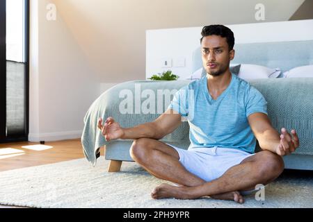 Uomo biraciale seduto sul pavimento in camera da letto, meditando Foto Stock