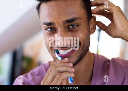 Felice uomo biraciale spazzolando i denti in bagno, sorridendo Foto Stock