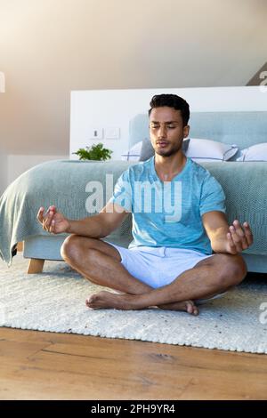 Uomo biraciale seduto sul pavimento in camera da letto, meditante, con spazio copia Foto Stock