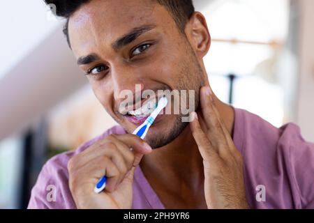 Felice uomo biraciale spazzolando i denti in bagno, sorridendo Foto Stock