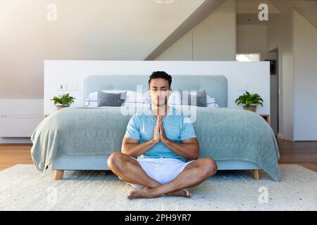 Uomo biraciale seduto sul pavimento in camera da letto, meditando Foto Stock