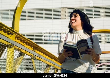 giovane donna latina, venezuelana, è ridente, ama leggere un libro mentre cammina per l'università, va con gli occhi chiusi e sorridente inte Foto Stock