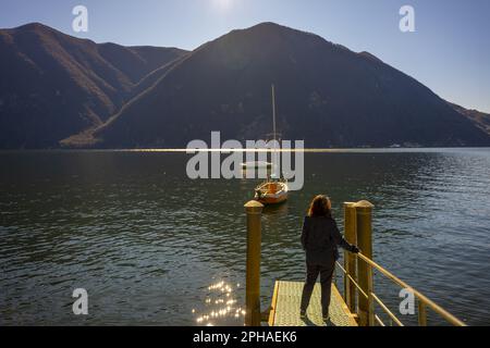 Donna in piedi su un molo con barca a vela sul Lago di Lugano e sulla catena montuosa in una giornata di sole con cielo limpido a Lugano, Ticino, Svizzera. Foto Stock