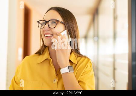 Allegro dipendente femminile ha conversazione telefonica in piedi in ufficio moderno, positivo giovane donna di affari felice di parlare con la collega sullo smartphone, ama chattare con gli amici durante una pausa Foto Stock