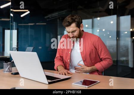 Un uomo in ufficio sul posto di lavoro è malato, ha un grave mal di stomaco, l'uomo d'affari ha avvelenato il cibo dal pranzo, è seduto al tavolo all'interno dell'edificio. Foto Stock