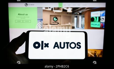 Persona che tiene uno smartphone con il logo della società olandese di commercio di automobili OLX Global B.V. sullo schermo di fronte al sito web. Messa a fuoco sul display del telefono.