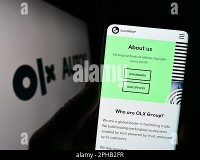 Persona in possesso di smartphone con il sito web della società olandese di commercio di automobili OLX Global B.V. sullo schermo di fronte al logo. Messa a fuoco al centro del display del telefono.