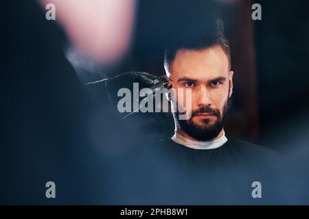 Giovane uomo barbuto seduto e di ottenere taglio di capelli in barbiere Foto Stock