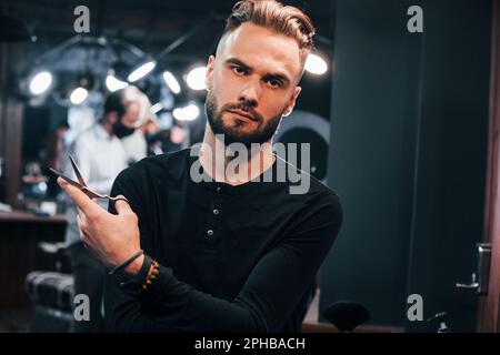 Giovane uomo bearded in piedi nel barbiere e tenendo le forbici Foto Stock