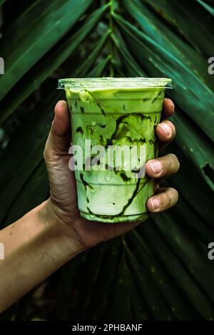 Tè ghiacciato verde matcha mescolato con cubetto di ghiaccio e latte in vetro latte con sfondo verde natura Foto Stock