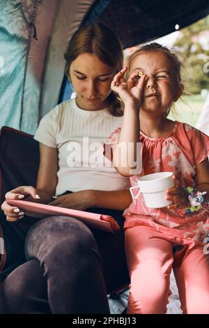 Due sorelle ragazze che trascorrono tempo in famiglia in una tenda in campeggio. I bambini che usano un tablet che giocano online durante le vacanze estive Foto Stock