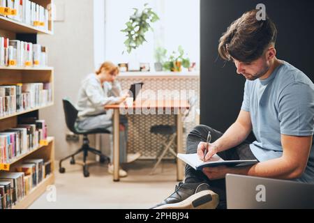 Studenti che imparano nella biblioteca universitaria. Giovane uomo che si prepara per il test prendendo appunti e usando il computer portatile. Ragazza e-book da tablet. Studia focalizzata Foto Stock