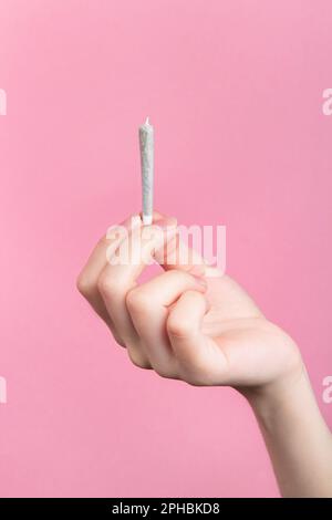 La mano di una donna tiene un'articolazione con la marijuana medica con le dita. Su sfondo rosa Foto Stock