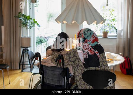 Vista posteriore della madre seduta con la figlia che ha paraplegia a casa Foto Stock