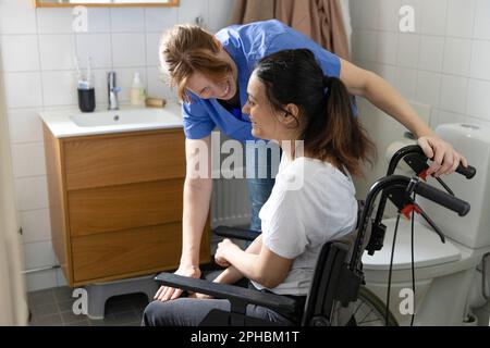 Felice donna caregiver che parla con una donna con paraplegia in bagno a casa Foto Stock