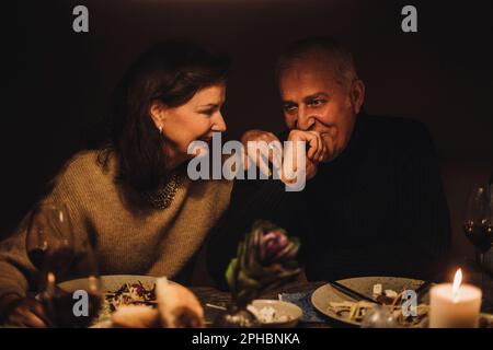 Donna anziana sorridente che parla con un uomo maturo con mano sul mento a cena Foto Stock