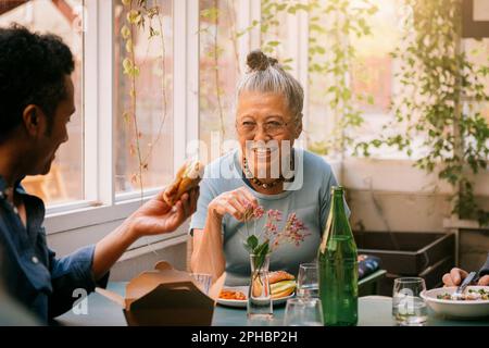 Donna anziana felice che si gode il cibo con un amico maschio mentre si siede al ristorante Foto Stock