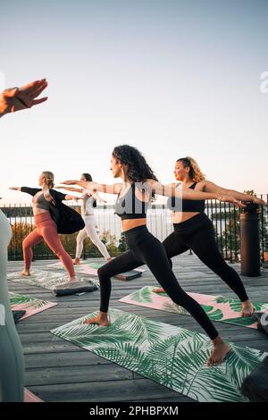 Le amiche che praticano il Guerriero 2 posano sul patio al tramonto Foto Stock