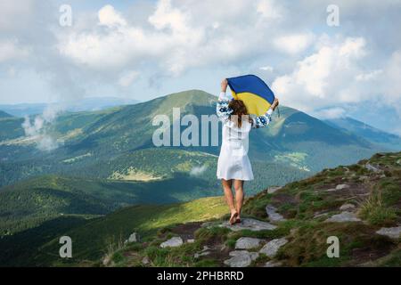 Una ragazza in un abito tradizionale ucraino ricamato si erge su una cima con una bandiera di Ucraina. La bandiera dell'Ucraina sulla cima di Hoverla in Ucraina Foto Stock