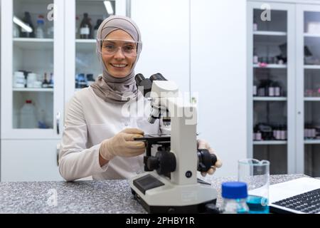 Ritratto di una giovane donna musulmana assistente di laboratorio, un medico seduto a un tavolo con un microscopio in un centro medico diagnostico. Guarda la fotocamera con un sorriso. Foto Stock