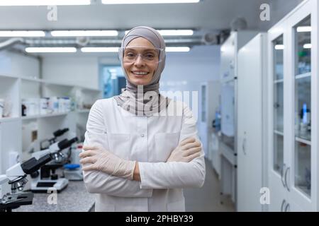 Ritratto di una giovane donna musulmana tecnico di laboratorio, scienziato, chimico, biologa che indossa un hijab in piedi in un centro di ricerca attraversando le braccia sul petto, guardando la macchina fotografica con un sorriso. Foto Stock