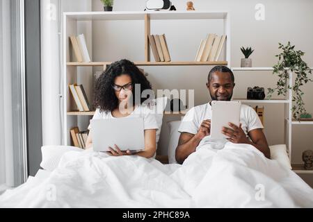 Vista frontale della coppia multiculturale innamorata che lavora su gadget moderni mentre si sdraiava sotto coperta a casa. Uomo sorridente che legge gli aggiornamenti sui social tramite tablet e donna serena che acquista online utilizzando un notebook. Foto Stock