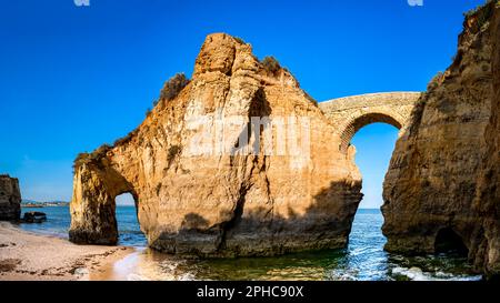 Vista a basso angolo delle rovine dell'antica passerella romana Ponte Romana de Lagos che collega due scogliere alla spiaggia Praia dos Estudantes, vicino Lagos. Foto Stock