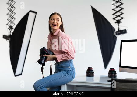 Entusiasta fotografo femminile seduto sul bordo del tavolo, tenendo la sua fotocamera DSLR e sorridendo, photostudio interni Foto Stock