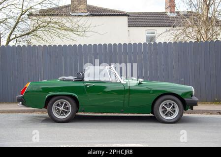 Green MG Midget parcheggiato da una recinzione di legno grigio con una casa dietro su una strada suburbana Foto Stock