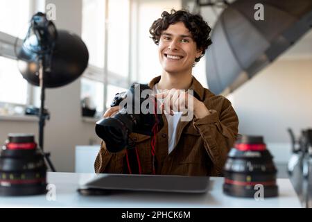 Allegro fotografo maschile seduto sul posto di lavoro in photostudio moderno, tenendo la fotocamera professionale e sorridendo Foto Stock