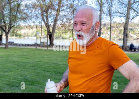 Un uomo anziano esausto sta catturando il respiro dopo essersi allenato nel parco pubblico della città vicino al fiume. Rinfrescando con acqua da una bottiglia di plastica. Foto Stock