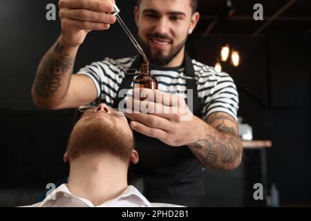 Parrucchiere con olio di barba presso il cliente in barbiere, primo piano. Servizio di rasatura professionale Foto Stock