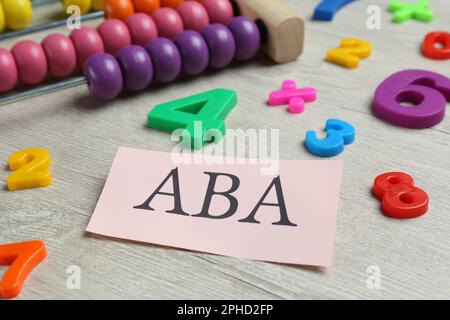 Foglio di carta con abbreviazione ABA (Applied Behavior Analysis), abaco e numeri colorati su tavola di legno bianco, primo piano Foto Stock