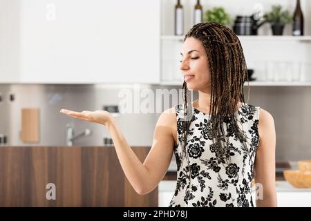 Splendida giovane donna con trecce e un vestito bianco e nero che si posa di fronte a uno sfondo moderno e luminoso della cucina, tenendo la mano come presenti Foto Stock