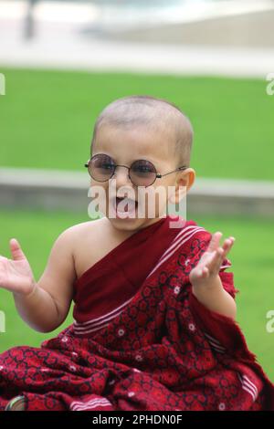 Un simpatico bambino calvo è vestito in avatar Monk, indossando uno scialle di maroon, un bicchiere da sole e dando un sorriso carino Foto Stock