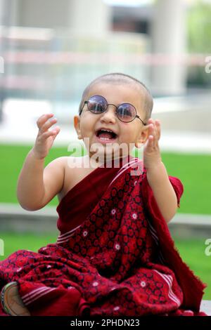 Un simpatico bambino calvo è vestito in avatar Monk, indossando uno scialle di maroon, un bicchiere da sole e dando un sorriso carino Foto Stock