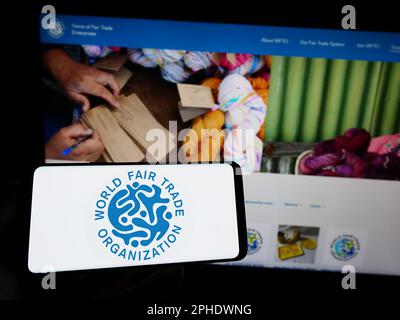 Persona che tiene in mano uno smartphone con il logo della World Fair Trade Organization (WFTO) sullo schermo di fronte al sito Web. Messa a fuoco sul display del telefono. Foto Stock