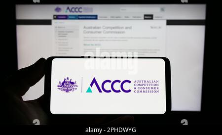 Persona che tiene in mano uno smartphone con il logo dell'Australian Competition e della Consumer Commission sullo schermo davanti al sito Web. Messa a fuoco sul display del telefono. Foto Stock