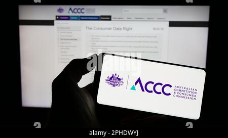 Persona che tiene il cellulare con il logo della concorrenza australiana e della commissione dei consumatori sullo schermo davanti alla pagina web. Messa a fuoco sul display del telefono. Foto Stock