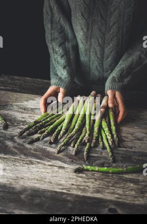 Persona irriconoscibile in maglione grigio in piedi al tavolo di legno con steli di asparagi freschi Foto Stock