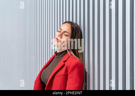 Giovane donna con mantello rosso e capelli biondi scuri appoggiati alla parete grigia con occhi chiusi nelle giornate di sole Foto Stock