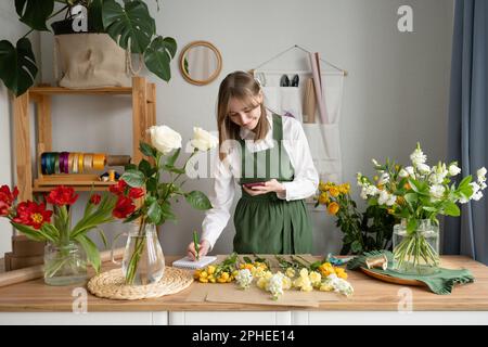 Donna allegra che indossa il verde generale e prendere appunti mentre si naviga smartphone e ricevere l'ordine in fioraio negozio con vari fiori Foto Stock