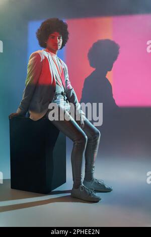 Corpo pieno di giovane modello maschile ispanico in tuta casual elegante seduta su cubo nero contro colorata illuminazione a strisce dal proiettore Foto Stock