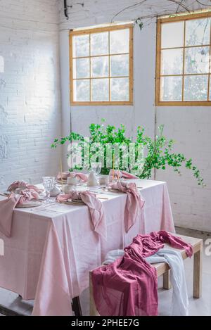 Tavolo in legno con tovaglia rosa e stoviglie collocato vicino alla panca coperta di stoffa contro piante in vaso e muro di mattoni bianchi Foto Stock