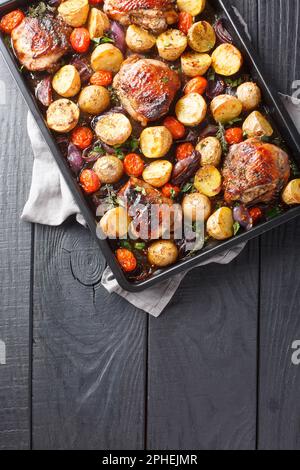 Deliziose cosce di pollo fatte in casa cotte con patate, aglio, cipolle e pomodori in aceto balsamico primo piano su una teglia nera su una ta di legno Foto Stock