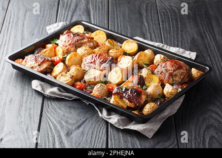 Cosce di pollo cotte con patate, aglio, cipolle e pomodori primo piano su una teglia nera su un tavolo di legno. orizzontale Foto Stock