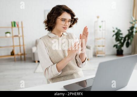 insegnante giovane in occhiali che mostra il gesto del linguaggio dei segni a due mani durante la lezione online, immagine stock Foto Stock