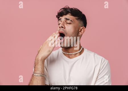 Giovane uomo elegante con occhi chiusi in collana che sbadigliava e copriva la bocca spalancata con la mano, mentre si trovava su un isolato dorso rosa Foto Stock