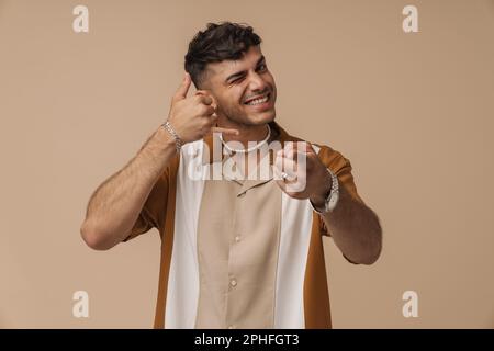 Giovane bell'uomo sorridente che fa un gesto telefonico con mano e dita, puntando e guardando la macchina fotografica, mentre si trova sopra isolato dorso marrone Foto Stock